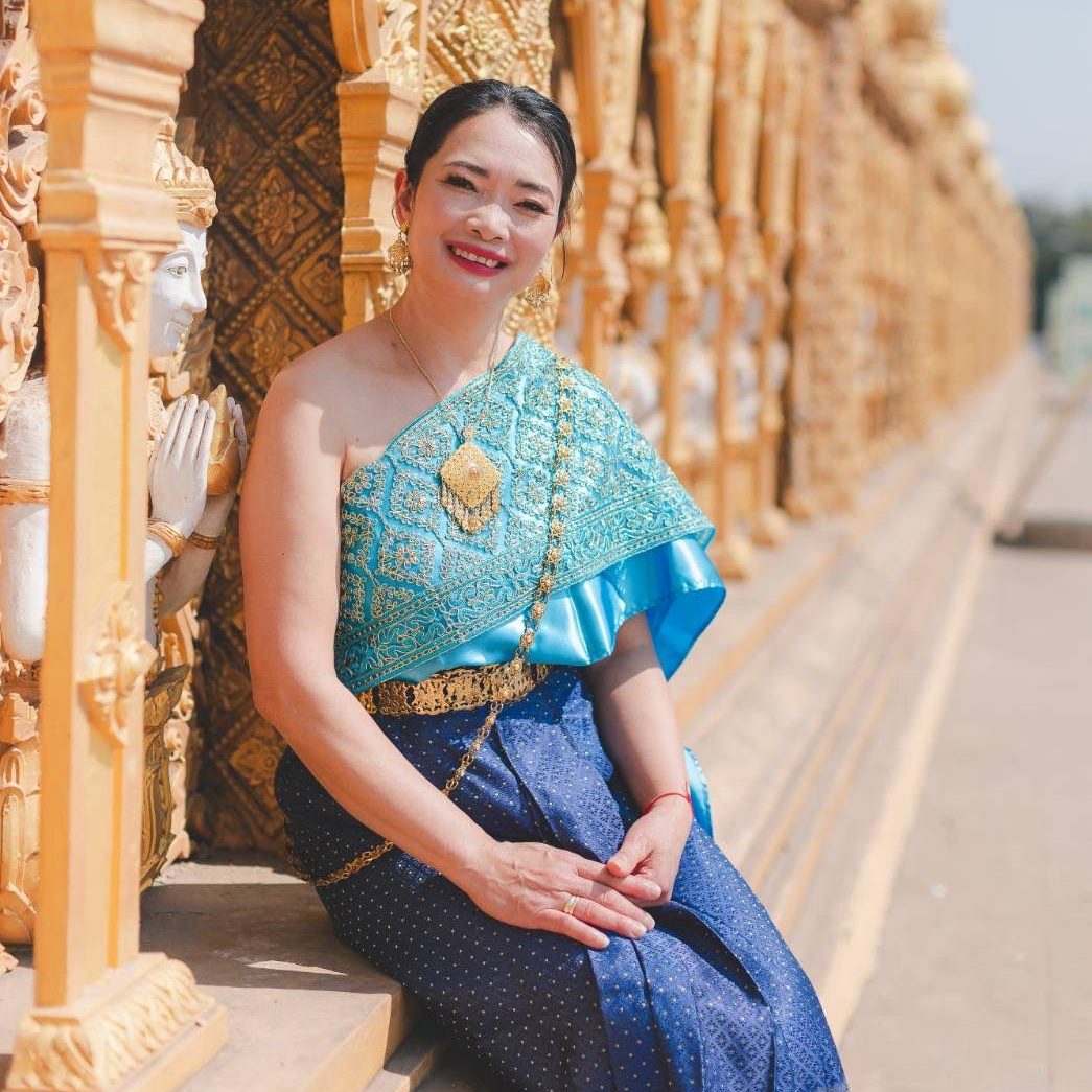 Auf dem Bild ist Gung zu sehen, welche lächelnd vor einem Tempel in Thailand in einem traditionellem thailändischen Gewand sitzt